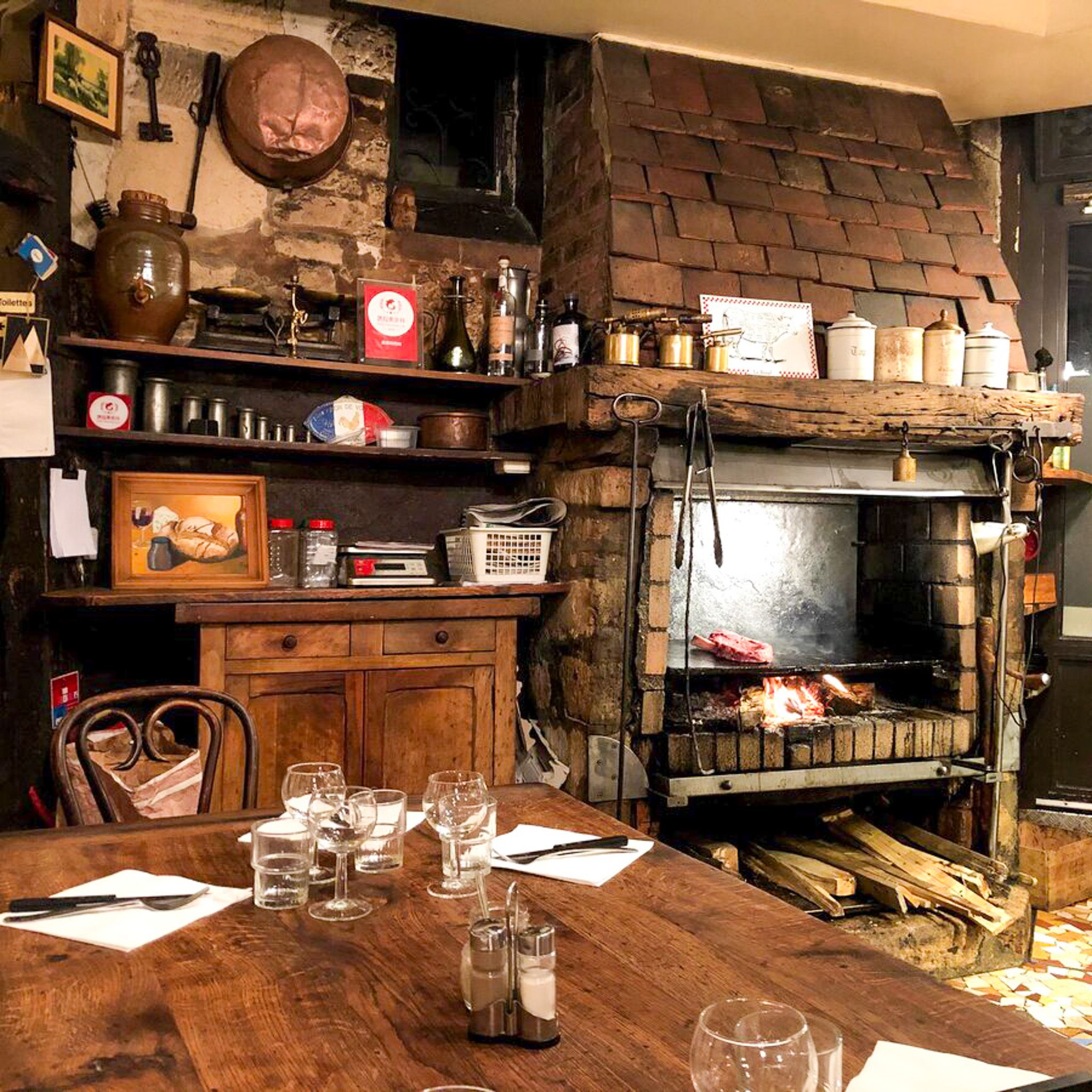 Rustic restaurant interior with steaks grilling in a large brick fireplace, and a wooden dining table set for guests in the foreground.
