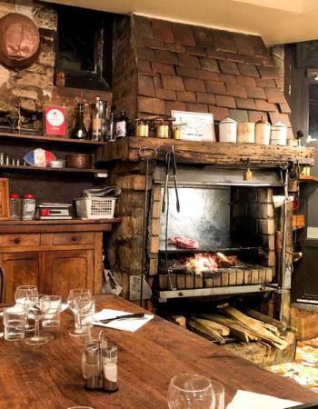 Rustic restaurant interior with steaks grilling in a large brick fireplace, and a wooden dining table set for guests in the foreground.