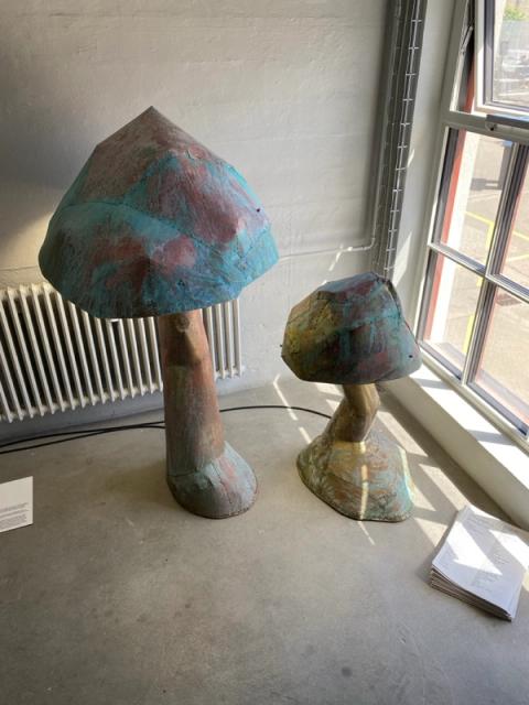 Two mushroom-shaped sculptures made of patinated copper stand indoors by a window.