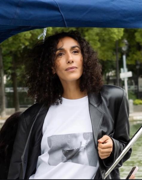 A woman with dark, curly hair in a black leather jacket and white t-shirt looks thoughtfully from a boat under a blue canopy.