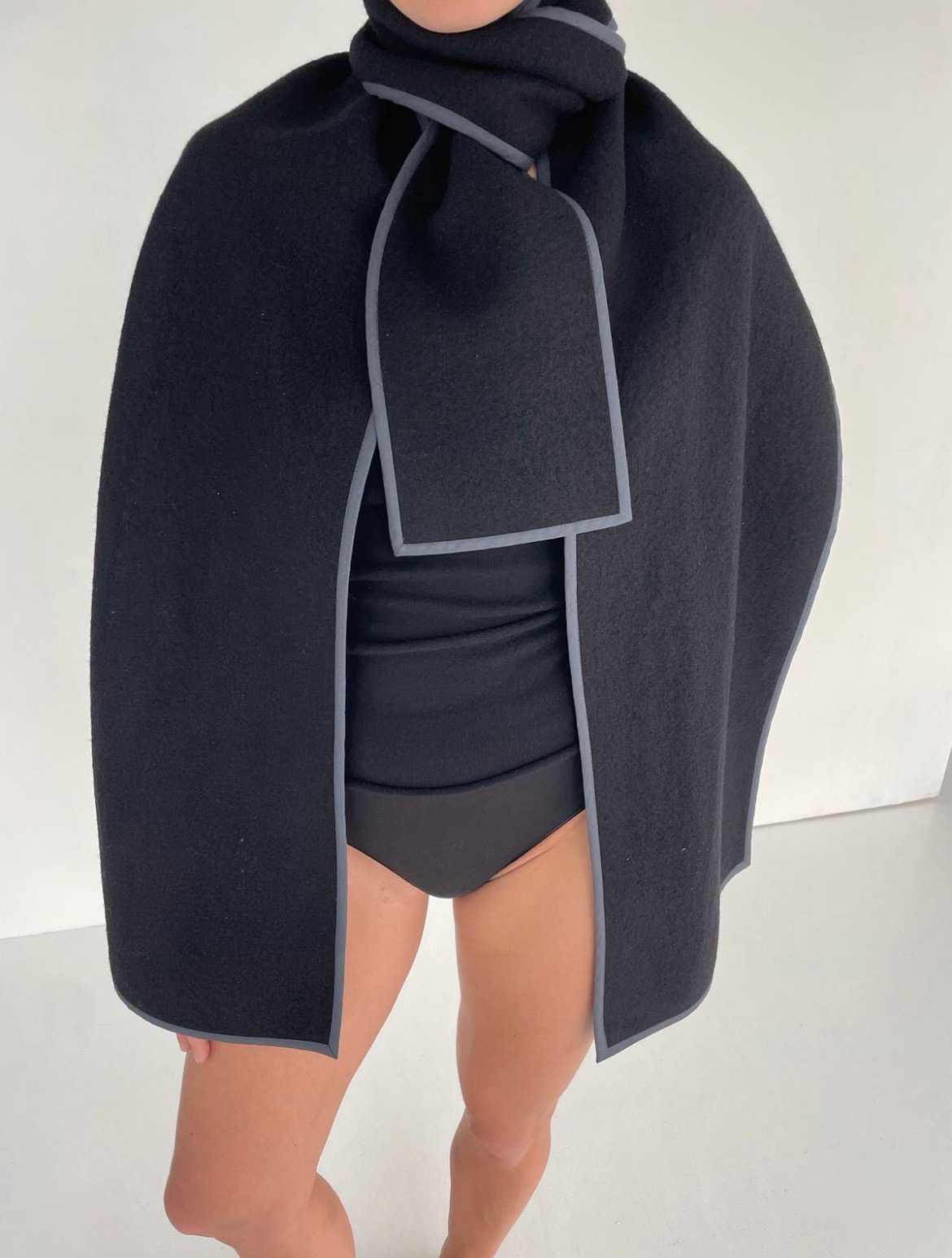 Person wearing a black felt cape and scarf with grey trim over black underwear and a top.