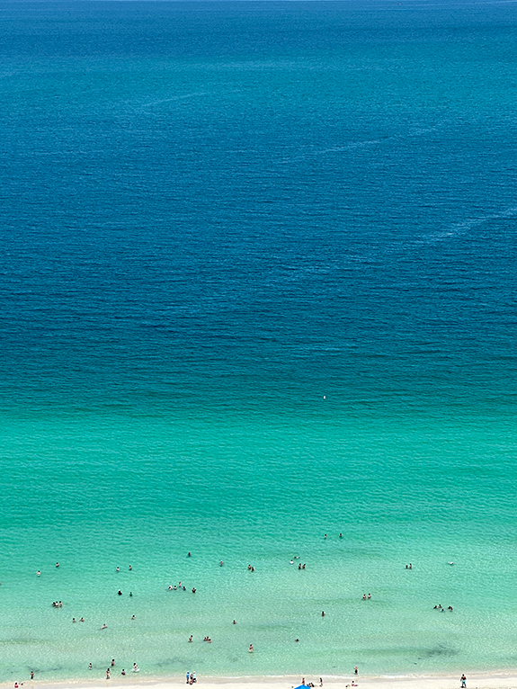 Aerial view of a beach with people swimming in clear turquoise water that transitions to a deep blue ocean.