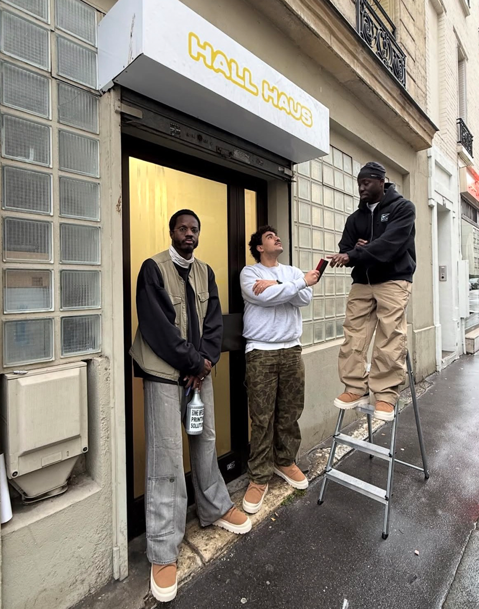 Three men stand outside a building with a "HALL HAUS" sign; one on a ladder reaches for an item from another, all wearing similar chunky shoes.