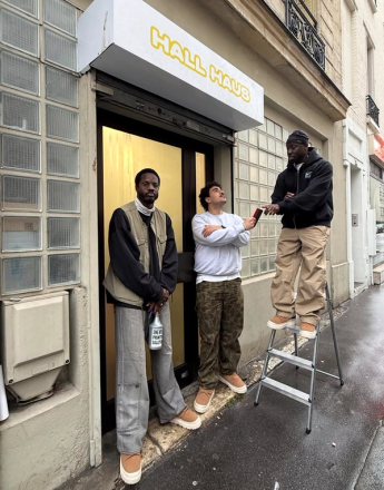 Three men stand outside a building with a "HALL HAUS" sign; one on a ladder reaches for an item from another, all wearing similar chunky shoes.
