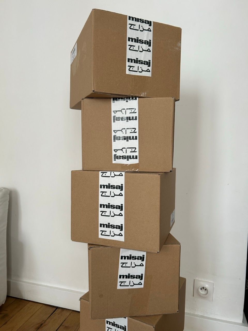 A tall stack of brown cardboard boxes sealed with white tape repeating "misaj" and Arabic script.