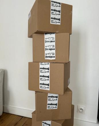 A tall stack of brown cardboard boxes sealed with white tape repeating "misaj" and Arabic script.