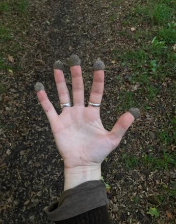 A hand with acorn caps on each fingertip and thumb, wearing two rings, against a leafy forest floor.