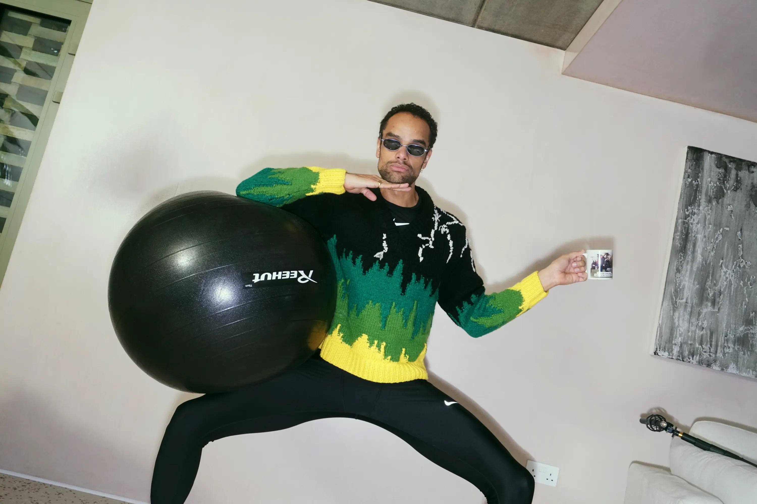 A man in sunglasses and a colorful sweater strikes a pose, holding a large black exercise ball and a white mug.