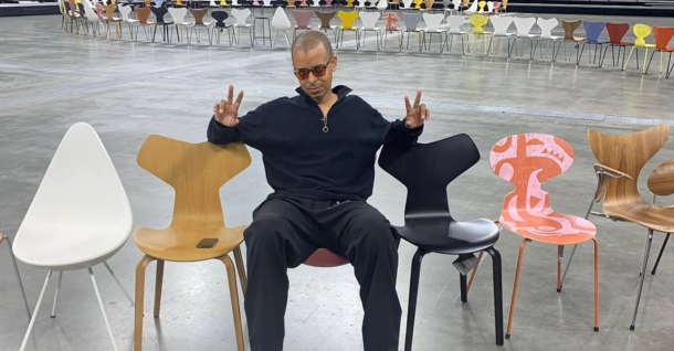 A man in sunglasses makes peace signs while seated among several modern chairs, with rows of similar chairs in the background.