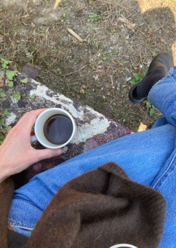 Overhead view of a person in jeans holding a coffee mug, seated on a stone ledge with a foot in a black shoe and dry ground visible.