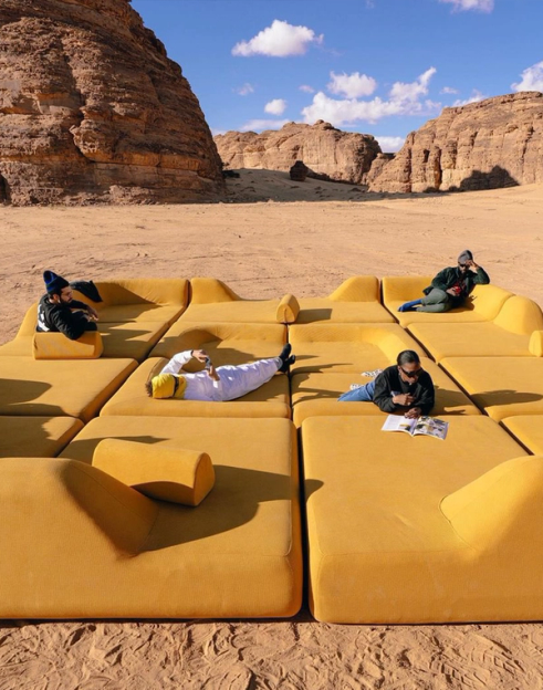 Four people lounge on a large, modular yellow sofa set up in a sandy desert surrounded by towering rock formations.
