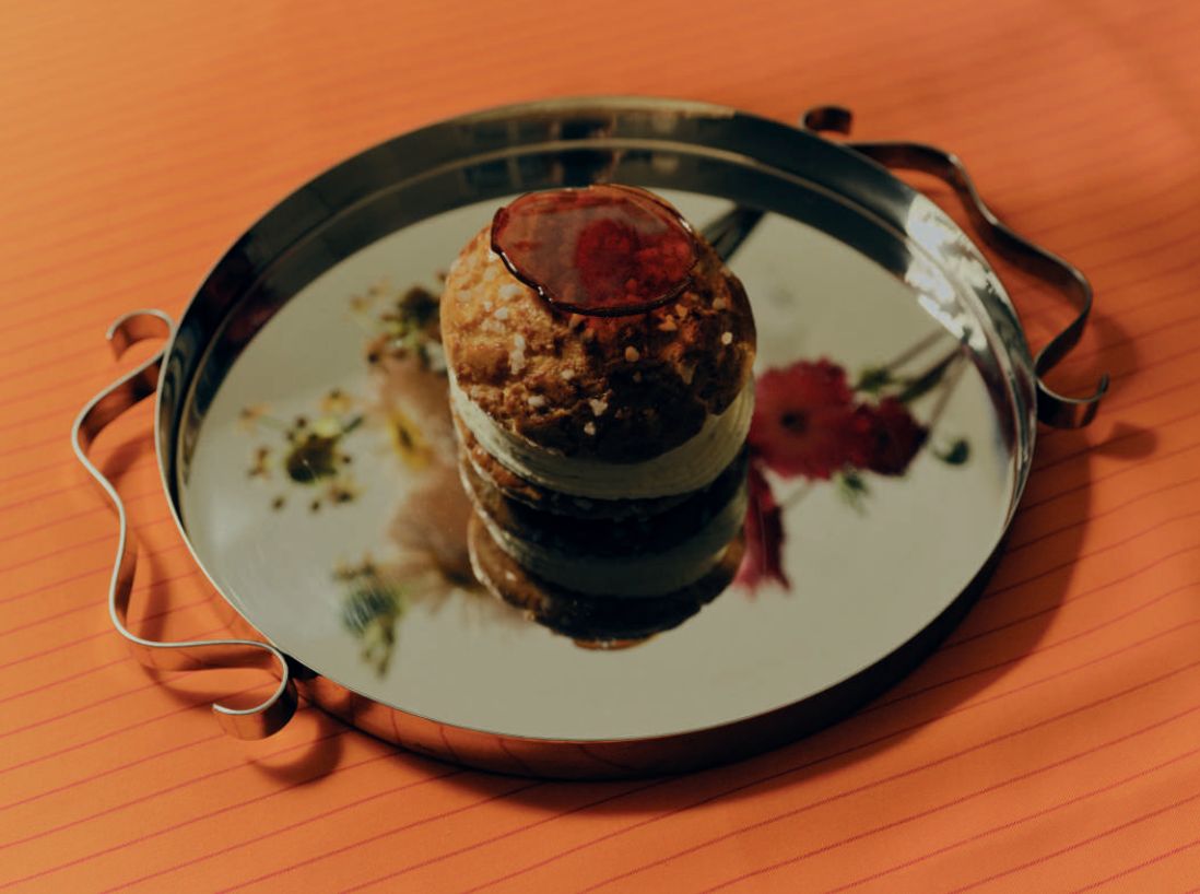 A round pastry with red glaze and sugar crystals sits on a reflective tray showing floral reflections.