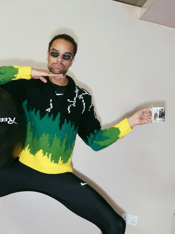 Man in sunglasses and a black, green, and yellow sweater striking a pose, holding a mug and a Reebok exercise ball.