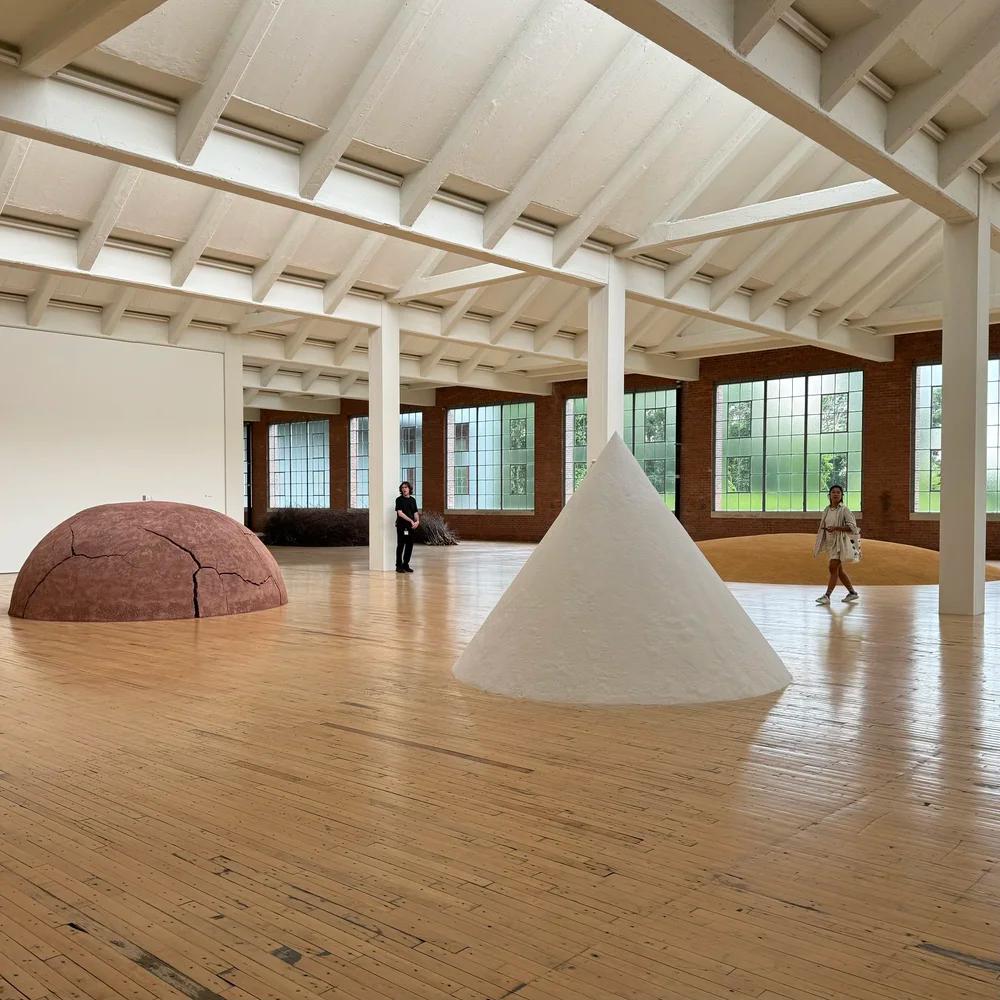 A large, bright gallery with wooden floors displays modern art: a reddish-brown cracked dome, a white cone, and a yellow gravel mound. Two visitors walk through the space.
