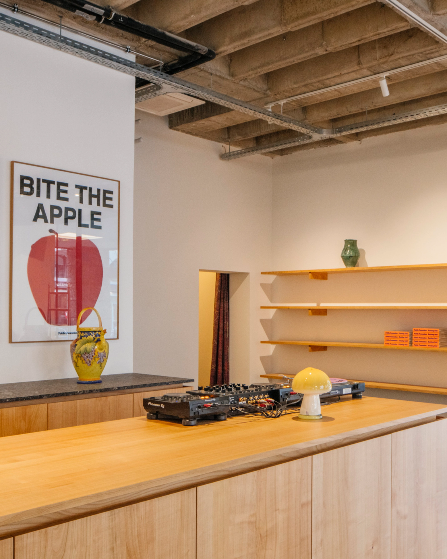 A modern room with a wooden counter, DJ equipment, a "BITE THE APPLE" poster, empty shelves, and an exposed concrete ceiling.