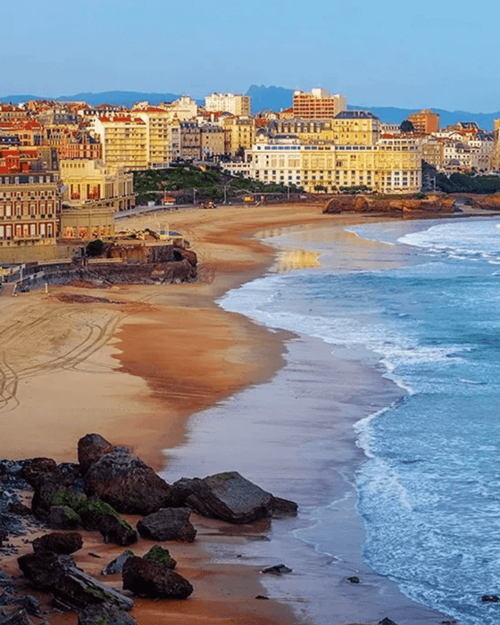 Coastal city buildings line a sandy beach with ocean waves and rocks.