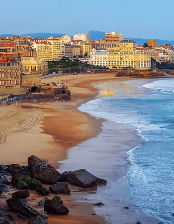 Coastal city buildings line a sandy beach with ocean waves and rocks.