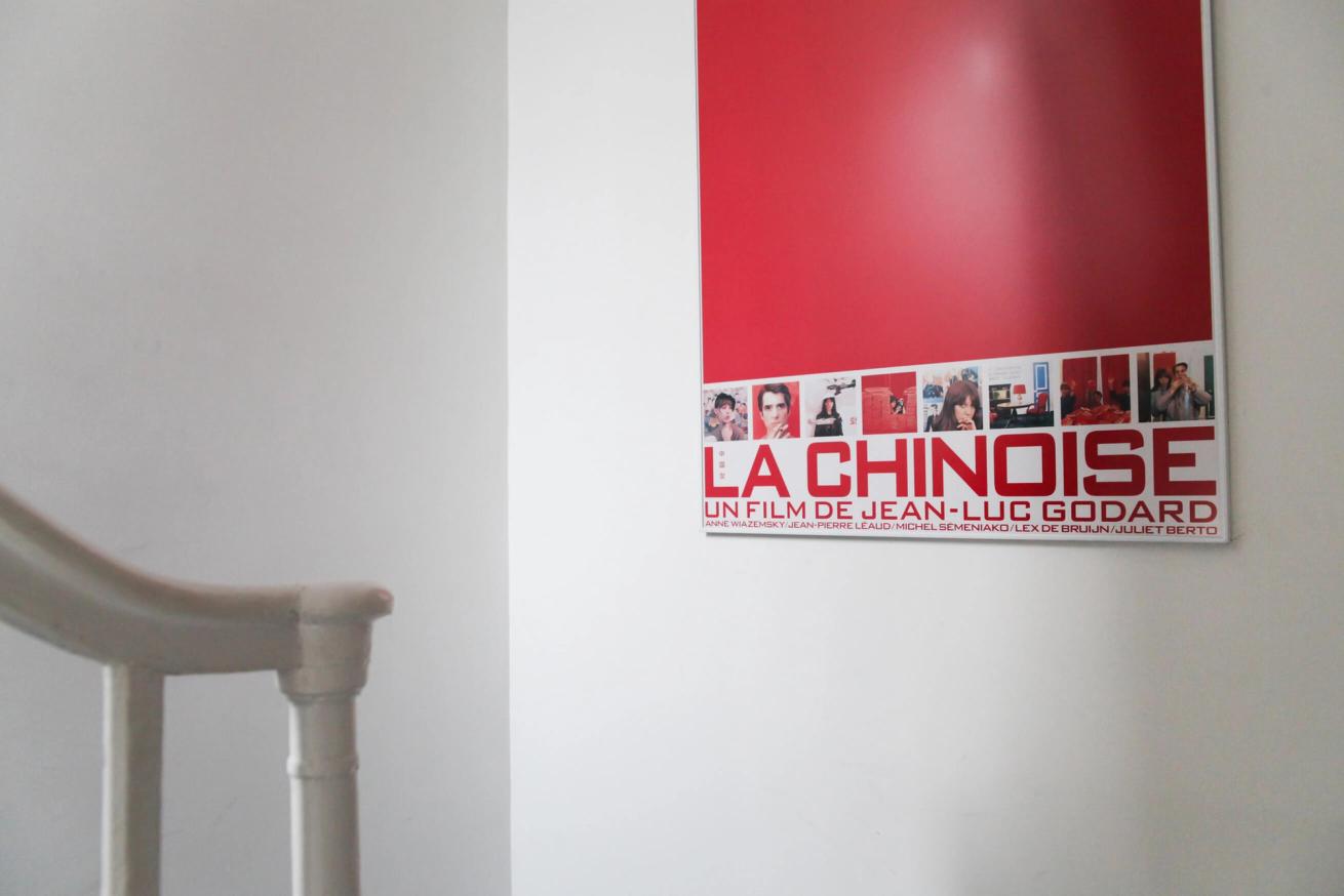 A red "La Chinoise" movie poster by Jean-Luc Godard hangs on a white wall, with a banister in the foreground.
