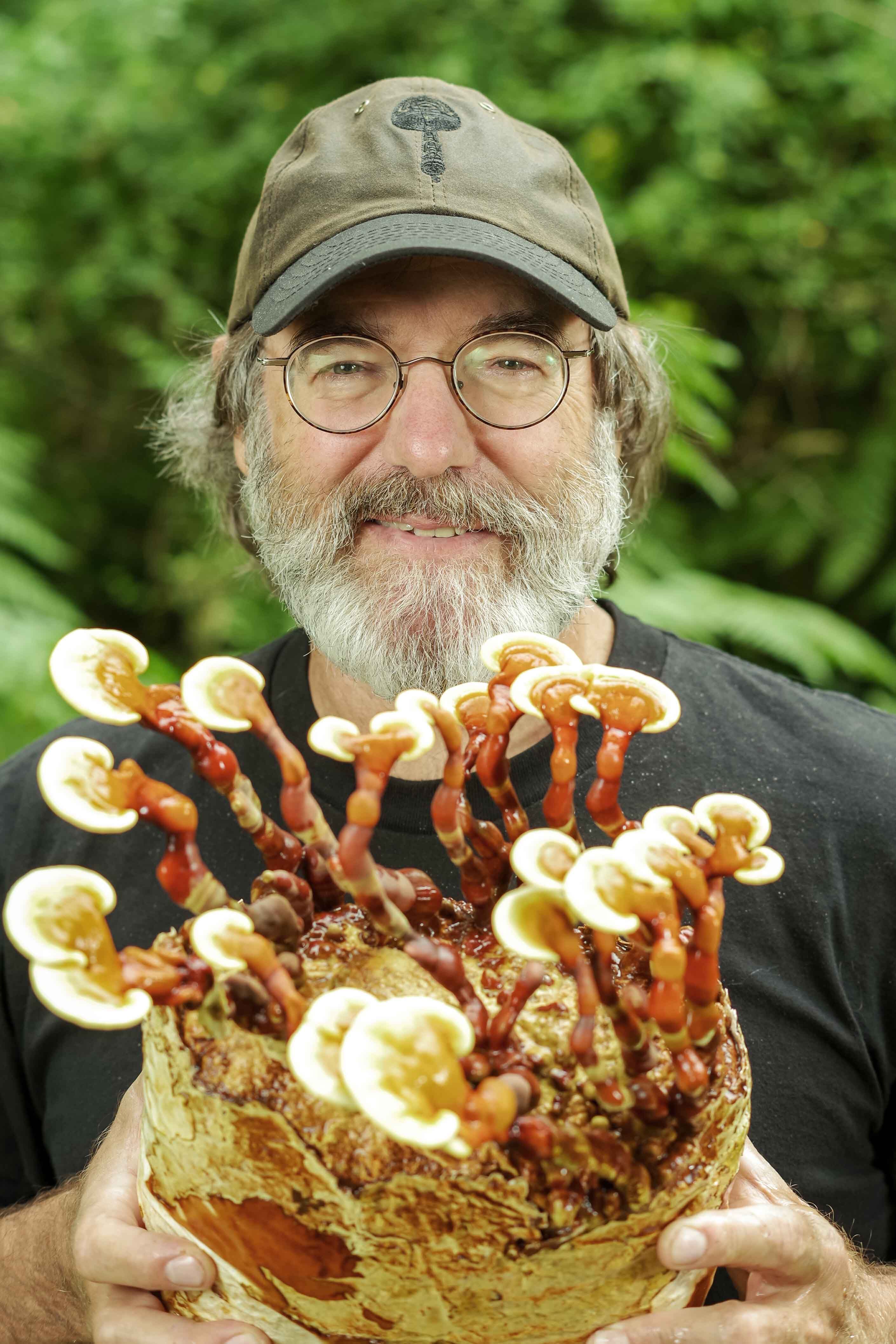 A smiling bearded man wearing glasses and a mushroom-logo cap holds a large cluster of glossy Reishi mushrooms.