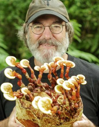 A smiling bearded man wearing glasses and a mushroom-logo cap holds a large cluster of glossy Reishi mushrooms.