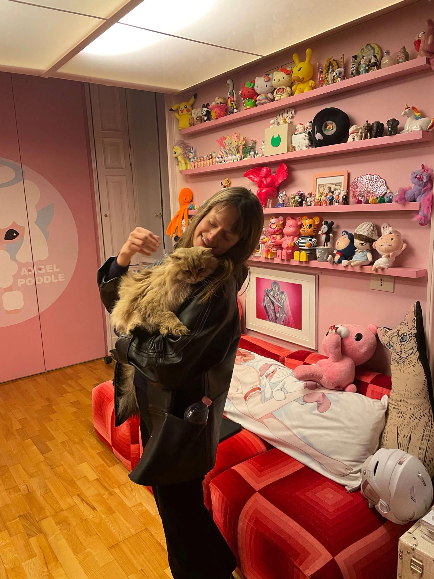 A smiling woman in a black jacket lovingly holds a fluffy cat in a pink room decorated with numerous pop culture collectibles on shelves.