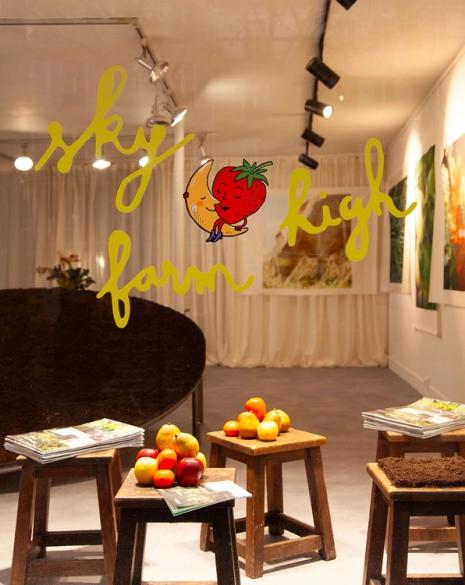 Shop window with "sky high farm" in yellow text and a cartoon moon hugging a strawberry, displaying fruit and magazines on wooden stools inside.