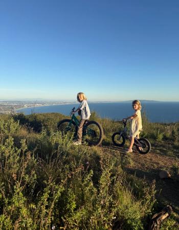 Two children with bikes on a grassy hill overlooking the ocean and a city.