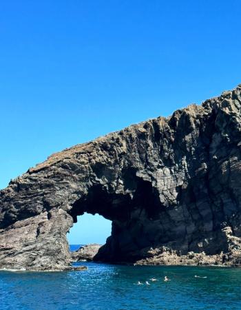 Natural rock arch over blue water with swimmers.