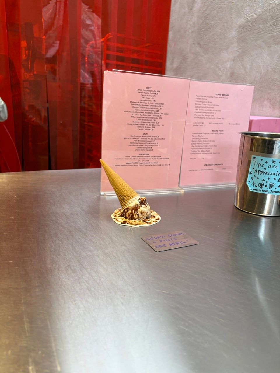 An overturned waffle cone with melted chocolate ice cream and sauce spilled on a metal counter, with a pink menu and tip jar in the background.