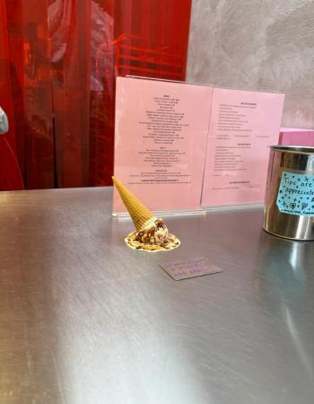 An overturned waffle cone with melted chocolate ice cream and sauce spilled on a metal counter, with a pink menu and tip jar in the background.