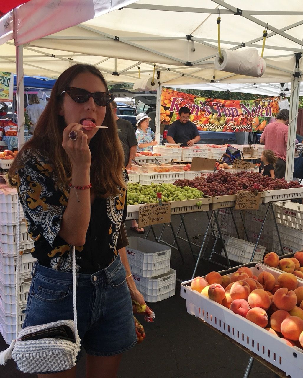 A woman in sunglasses licks a pink popsicle at a bustling farmer's market with tables of fresh produce.