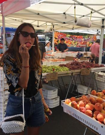 A woman in sunglasses licks a pink popsicle at a bustling farmer's market with tables of fresh produce.