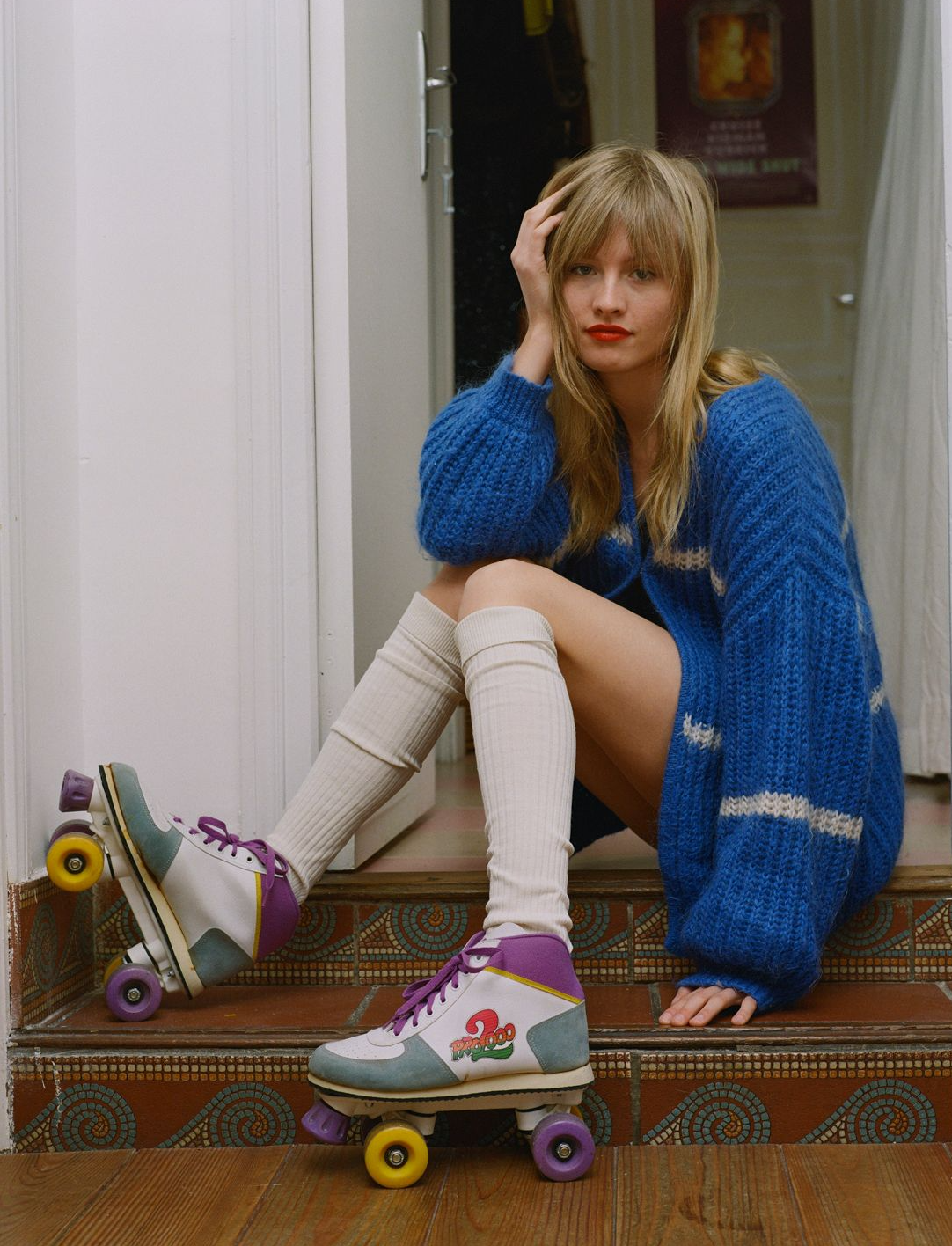 A blonde woman in a blue knit cardigan, cream knee-high socks, and retro roller skates sits on tiled steps.