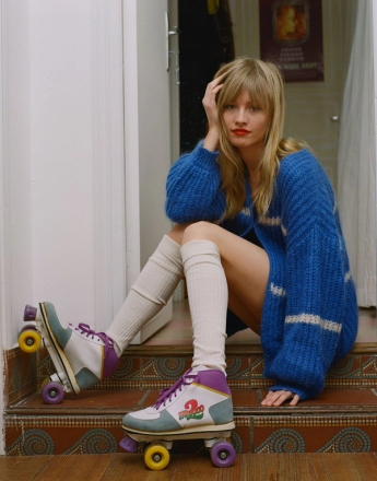 A blonde woman in a blue knit cardigan, cream knee-high socks, and retro roller skates sits on tiled steps.