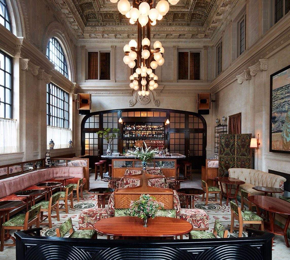 An opulent bar and lounge features a grand chandelier, high ornate ceiling, arched windows, and diverse plush seating arrangements.
