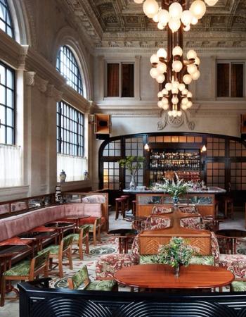 An opulent bar and lounge features a grand chandelier, high ornate ceiling, arched windows, and diverse plush seating arrangements.