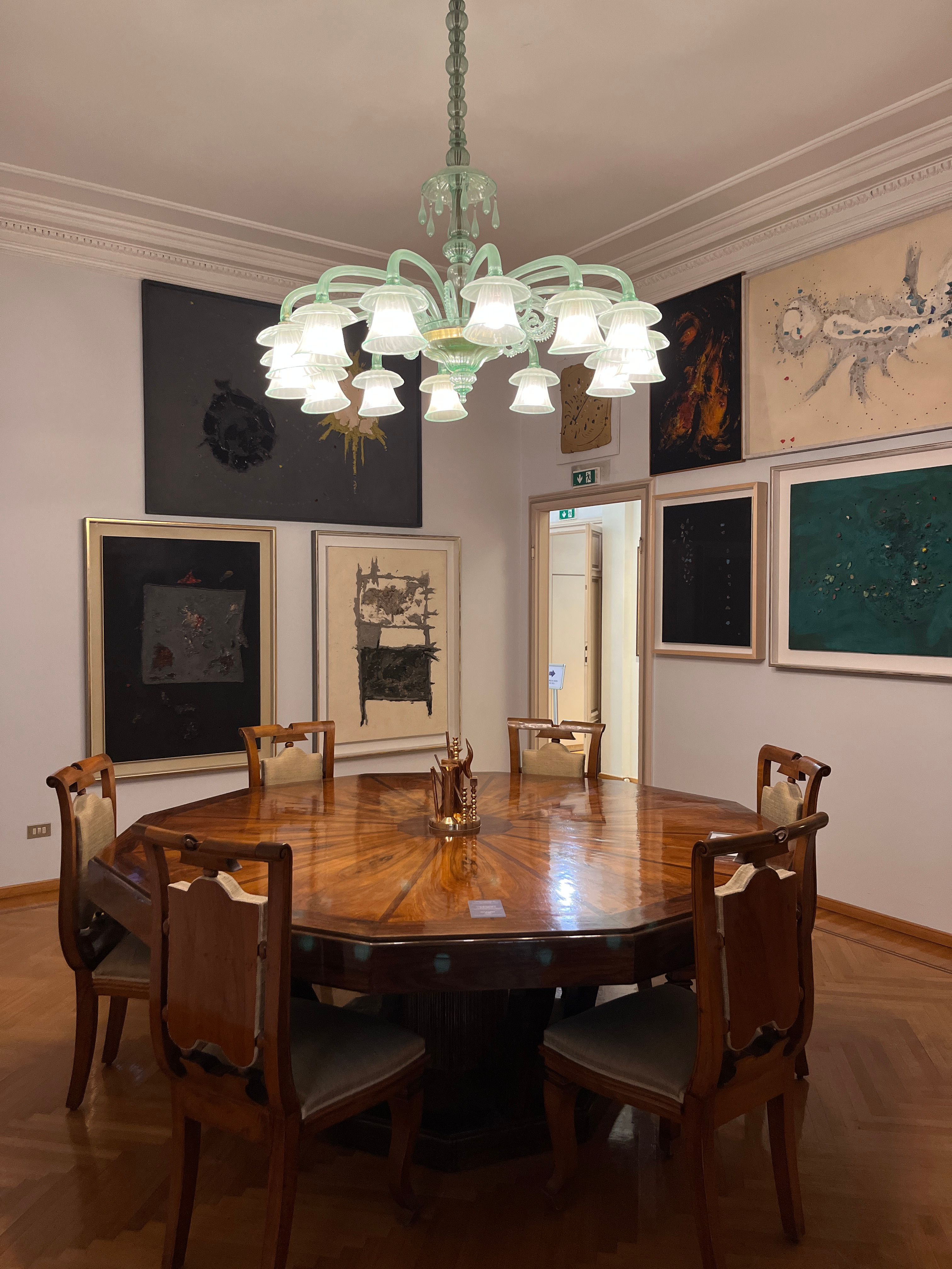 An octagonal wooden dining table surrounded by chairs under a green glass chandelier, with multiple abstract artworks on the walls.