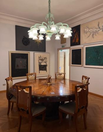 An octagonal wooden dining table surrounded by chairs under a green glass chandelier, with multiple abstract artworks on the walls.