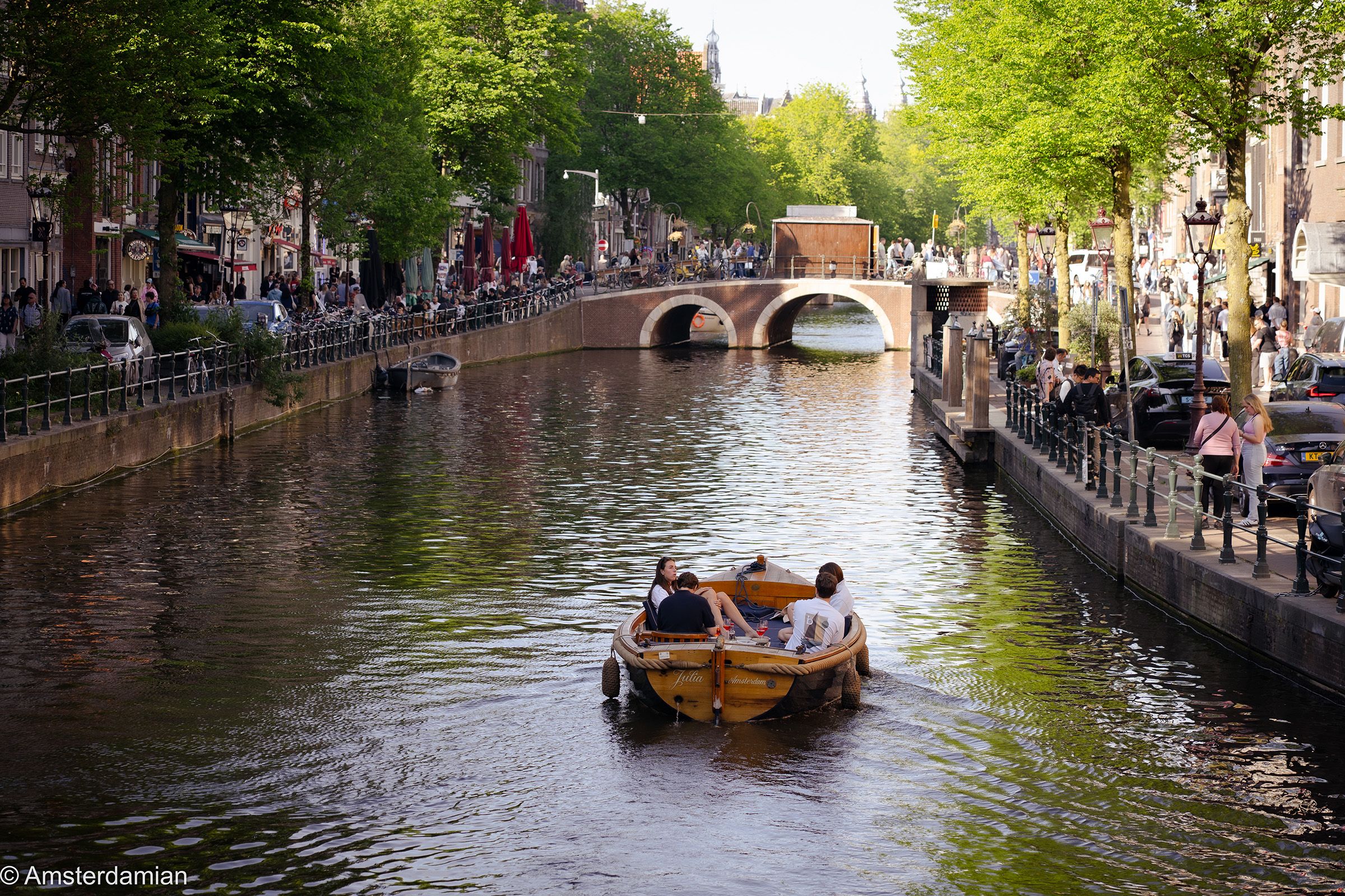 A boat carrying people cruises a city canal, flanked by buildings, green trees, and a distant bridge.