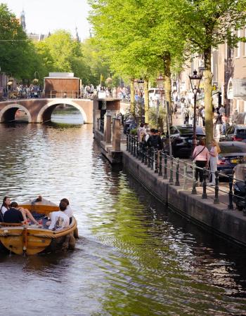 A boat carrying people cruises a city canal, flanked by buildings, green trees, and a distant bridge.
