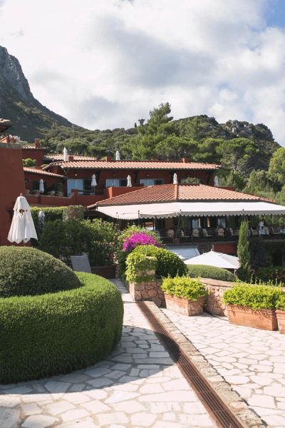 Terracotta-roofed buildings nestled in lush gardens with a stone pathway, backed by mountains under a cloudy sky.