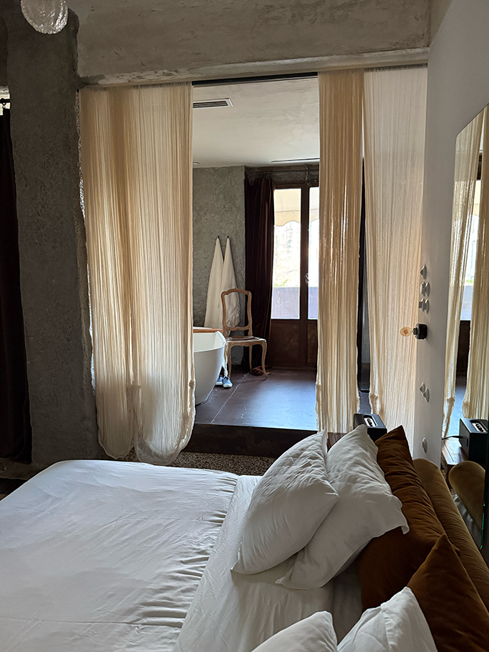 A bedroom with a white bed and sheer curtains opening to a bathroom with a freestanding tub.