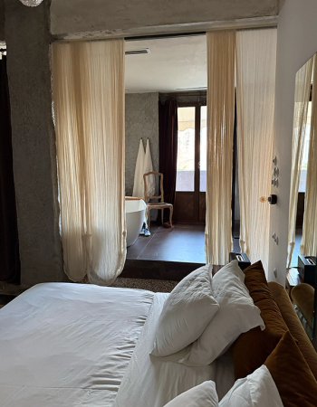 A bedroom with a white bed and sheer curtains opening to a bathroom with a freestanding tub.