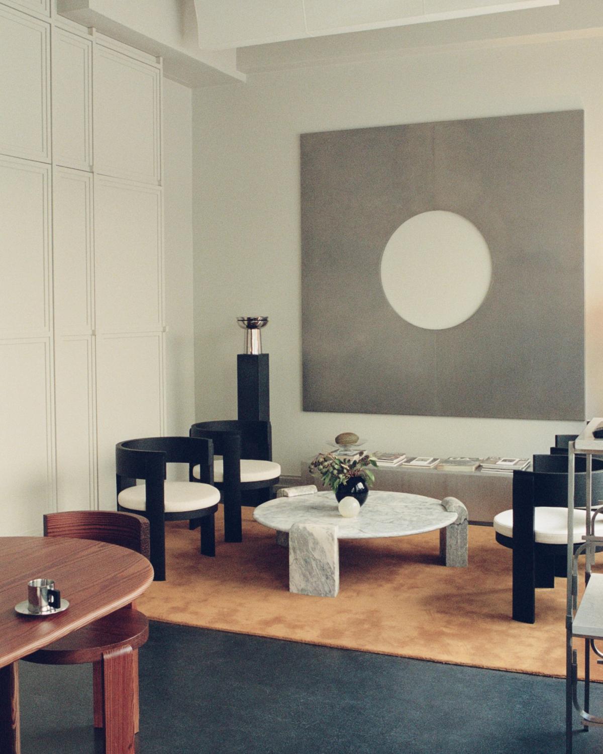 A modern living room with a marble coffee table, black chairs, an orange rug, and a large grey wall art with a white circle.