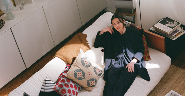 A woman in a dark outfit lounges on a white futon in a sunlit room, looking at the camera.