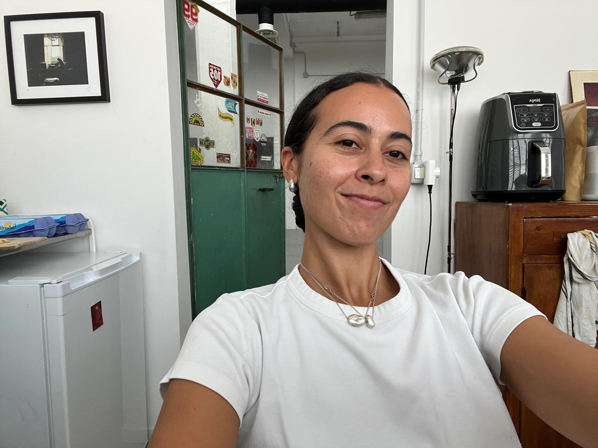 A young woman with dark hair in a white t-shirt and silver necklaces smiles gently, with a green door and an air fryer in the background.