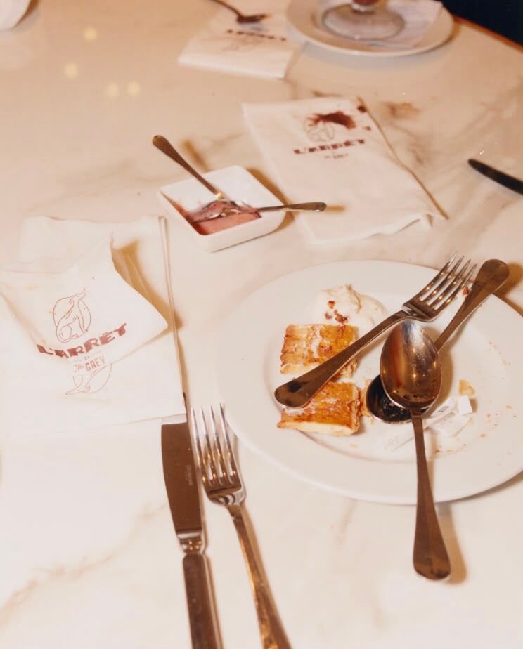 A messy table after a meal with leftover food, used cutlery, and stained napkins on a marble surface.