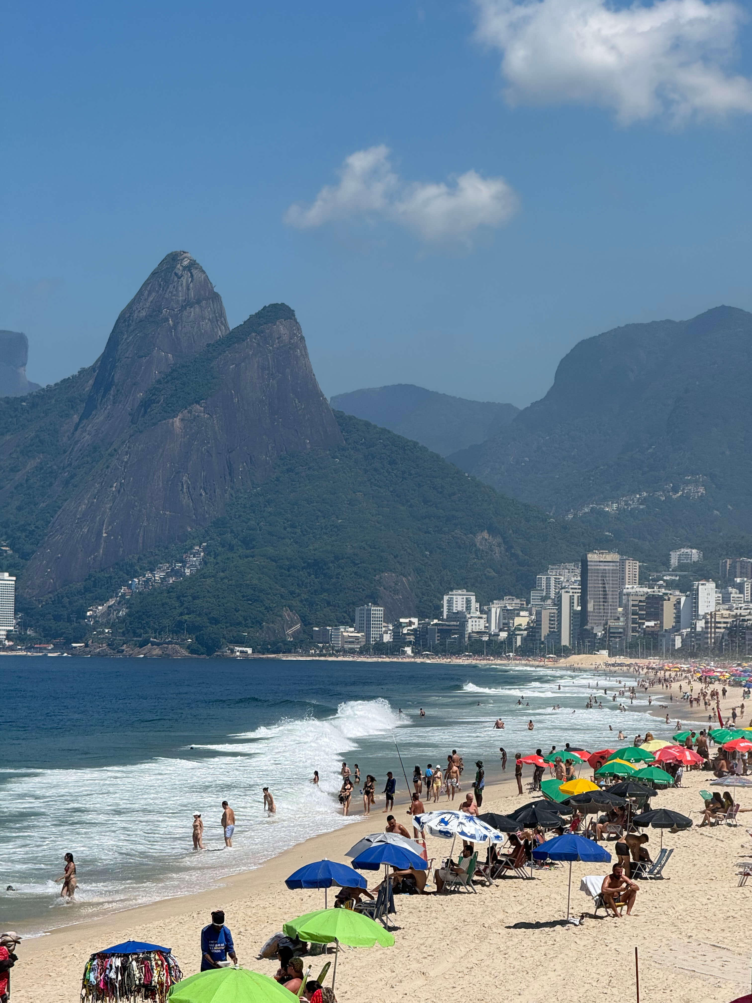 Sunny, crowded beach with people, colorful umbrellas, and waves, backed by prominent mountains and a city skyline.