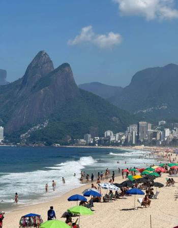 Sunny, crowded beach with people, colorful umbrellas, and waves, backed by prominent mountains and a city skyline.