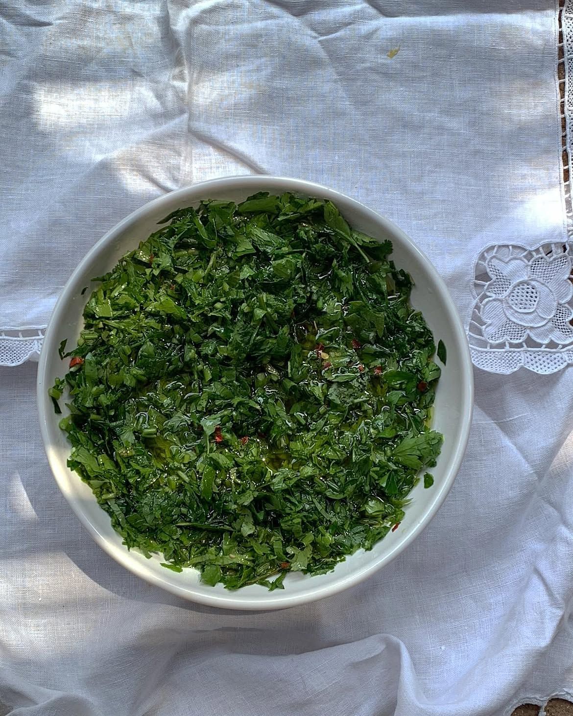 A white bowl of vibrant green chopped herbs with olive oil and red flecks on a white lace tablecloth.